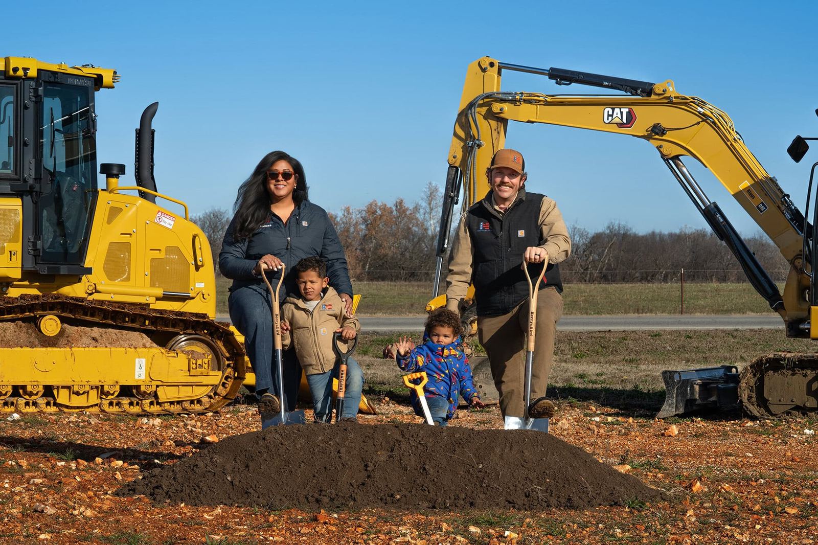 The Nash family - husband, wife, and their two children - holding shovels at a residential groundbreaking with CAT construction equipment behind them in Northwest Arkansas